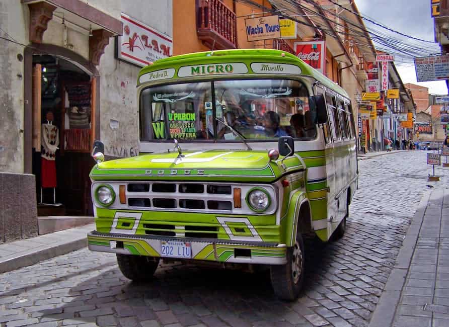 Vibrant green public bus navigating a bustling cobbled street in La Paz, Bolivia, with street vendors and shops.