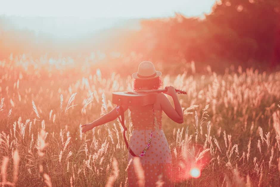 Woman in a field at sunset holding a guitar, embracing tranquility and freedom.